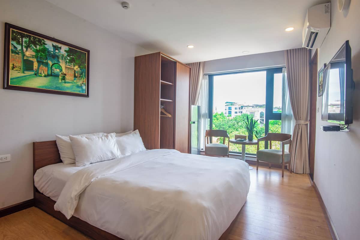A hotel room with a large bed, a wardrobe, a small table with chairs, and a large window offering a view of buildings and greenery.