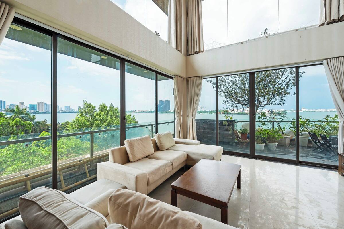 A spacious living room with large windows offering a city and water view, a beige sofa, and a wooden coffee table.