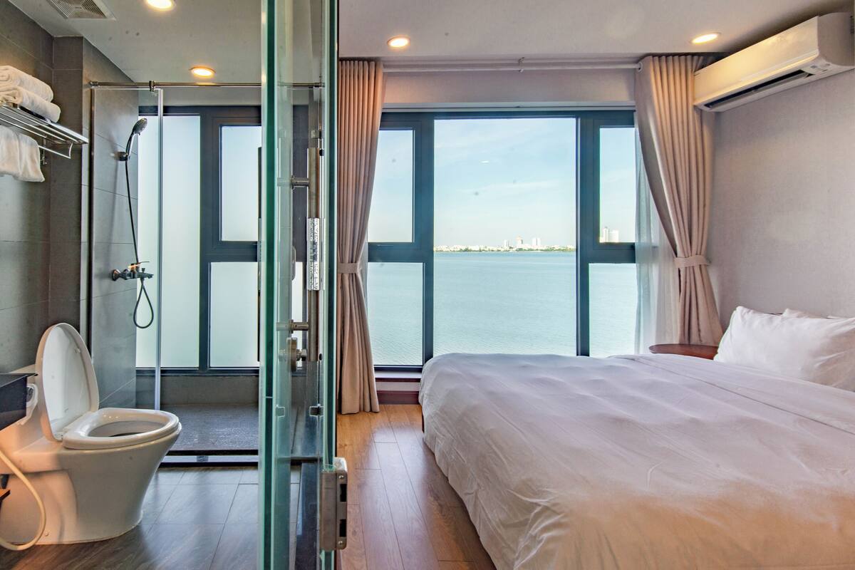A hotel room with a large bed, a glass-enclosed shower, a toilet, and a view of the water.