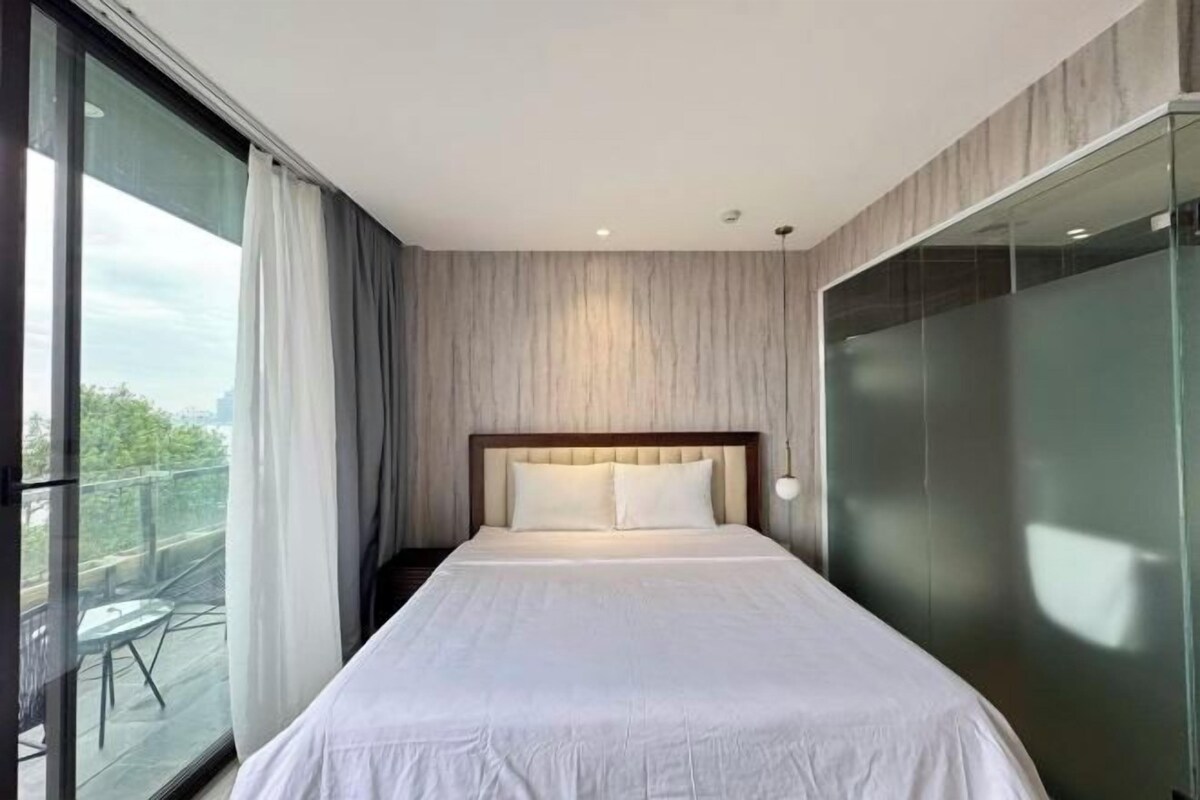 A modern hotel room with a large bed, a glass wall, and a balcony with outdoor furniture.