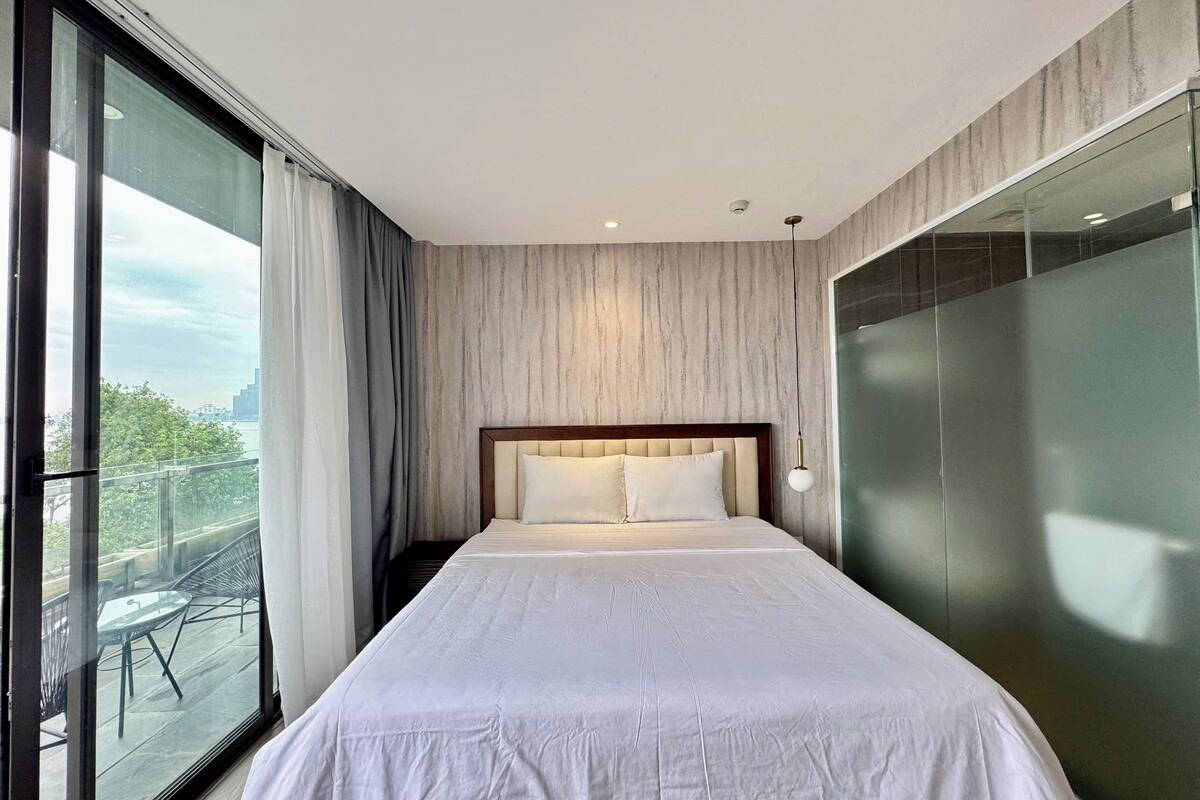 A modern hotel room with a large bed, a view of the city through floor-to-ceiling windows, and a balcony with outdoor furniture.