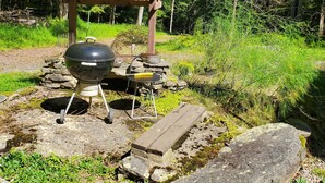A barbecue setup with a grill, a table, and a wooden shelter in a forested area. - Shambhala Mountain Retreat: A chalet surrounded by forest. Relax & unwind! (Napanoch)