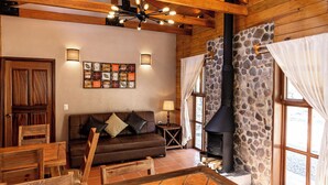 A cozy living room with a stone fireplace, wooden beams, a brown leather sofa, and a modern light fixture. - Roca Venados (Mascota)