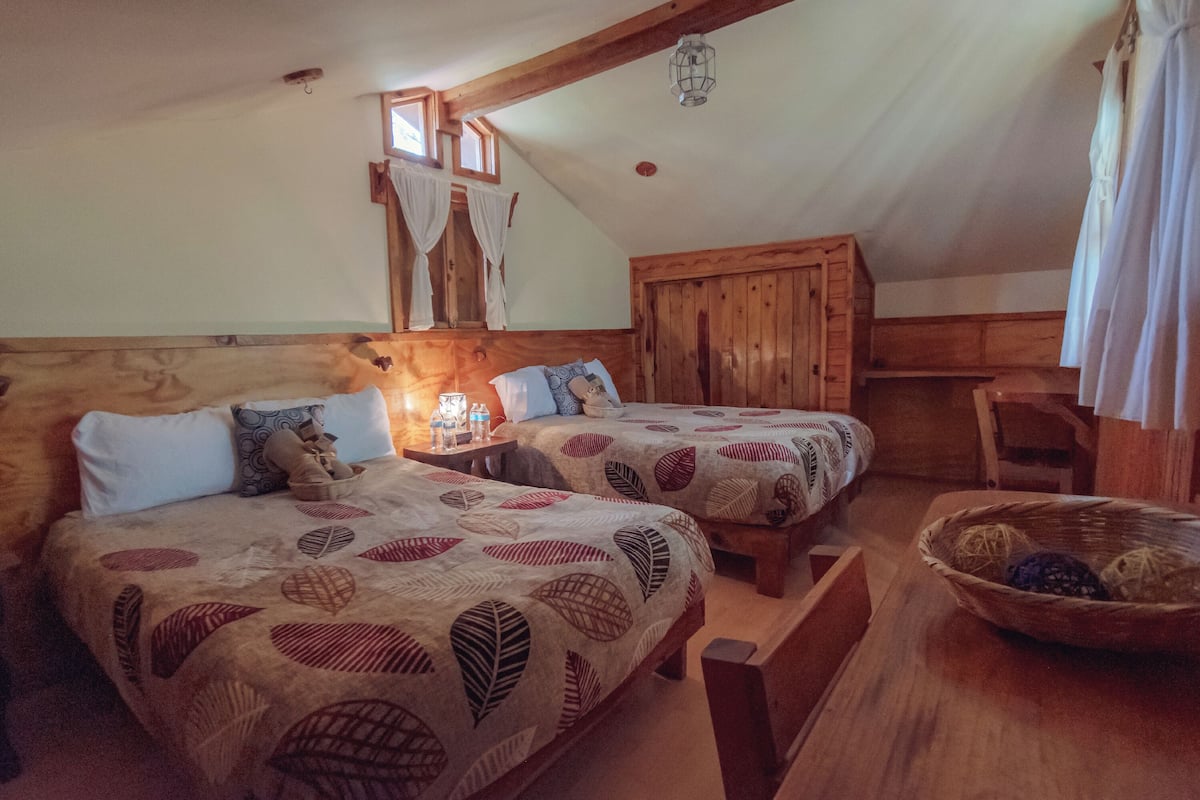 A cozy room with two beds, a wooden bench, a table with a basket, and a window with curtains.
