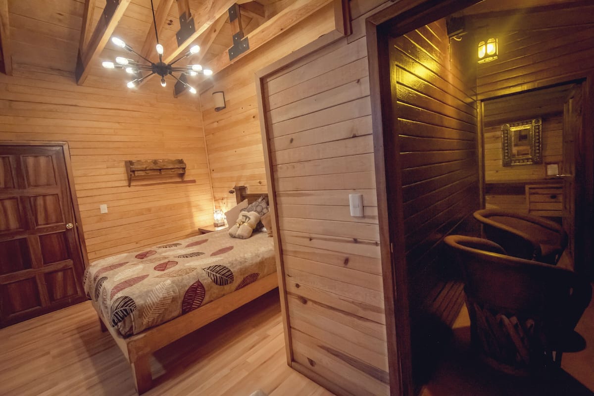 A wooden cabin room with a bed, a bench, a small table, and a mirror.