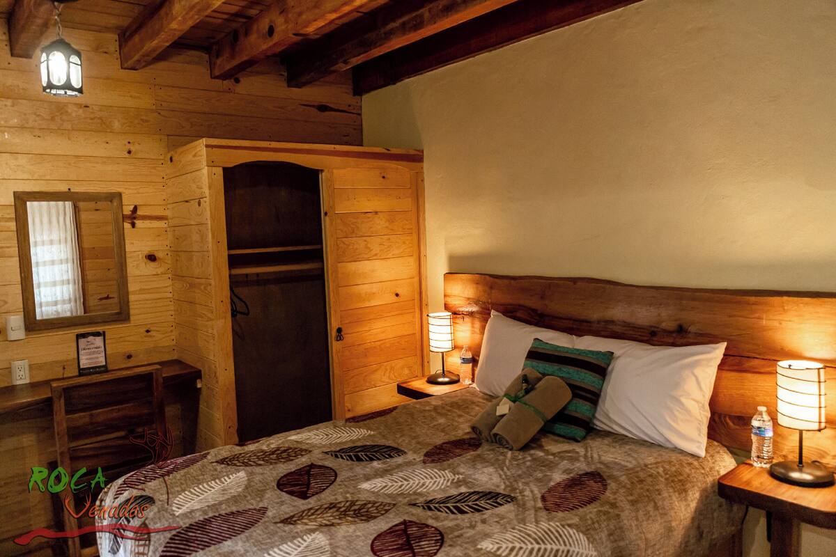 A wooden cabin-style room with a bed, two bedside tables with lamps, a nightstand, a mirror, and a door.