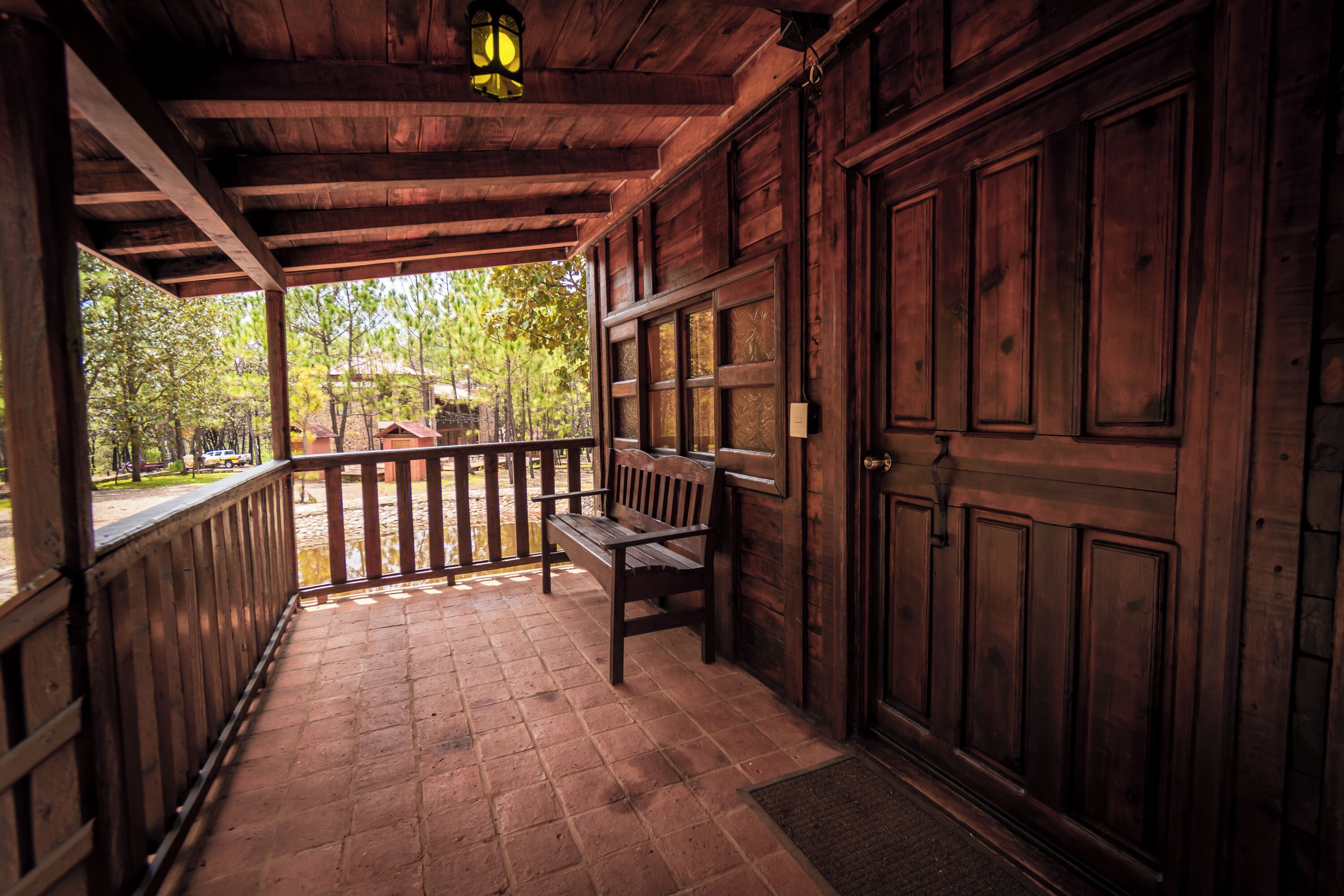 A wooden porch with a bench, a door, and a view of trees and a building in the background.