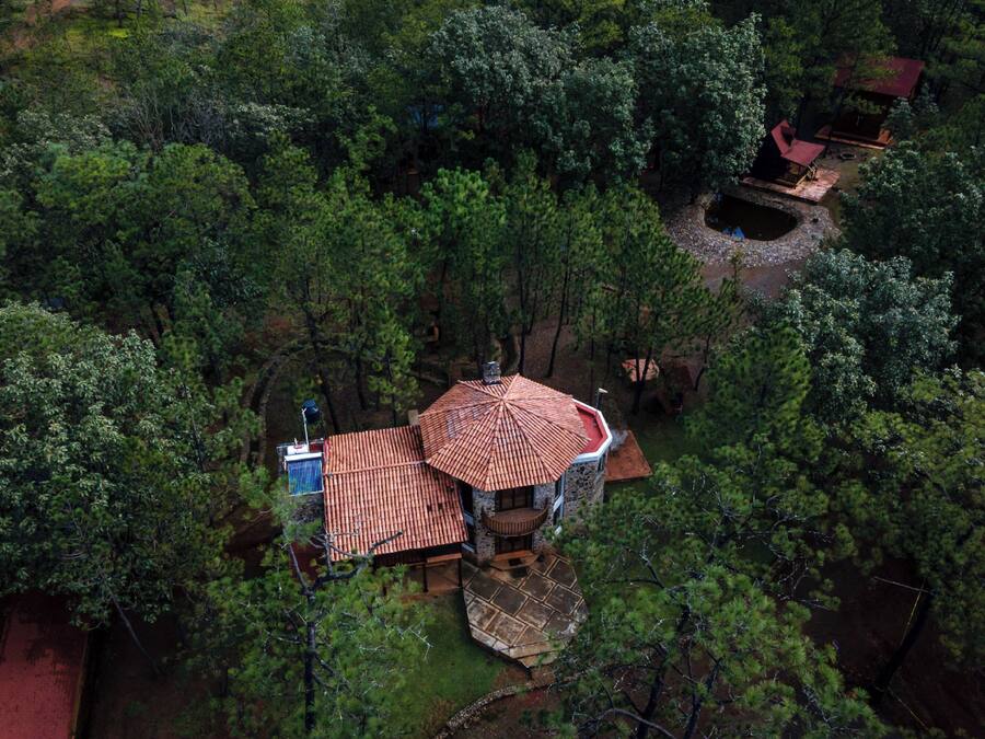 An aerial view of a secluded cabin surrounded by dense forest, featuring a central building with a red-tiled roof and a small pond nearby.