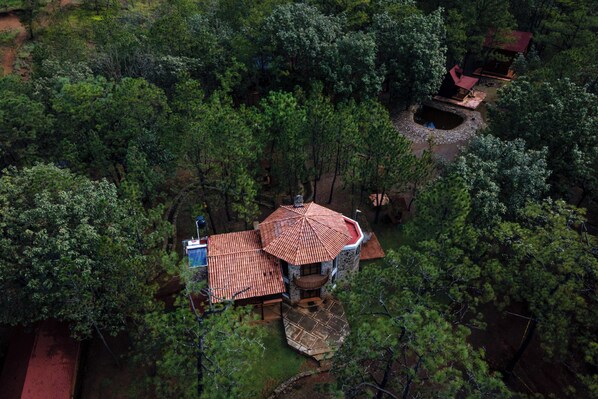 An aerial view of a secluded cabin surrounded by dense forest, featuring a central building with a red-tiled roof and a small pond nearby.