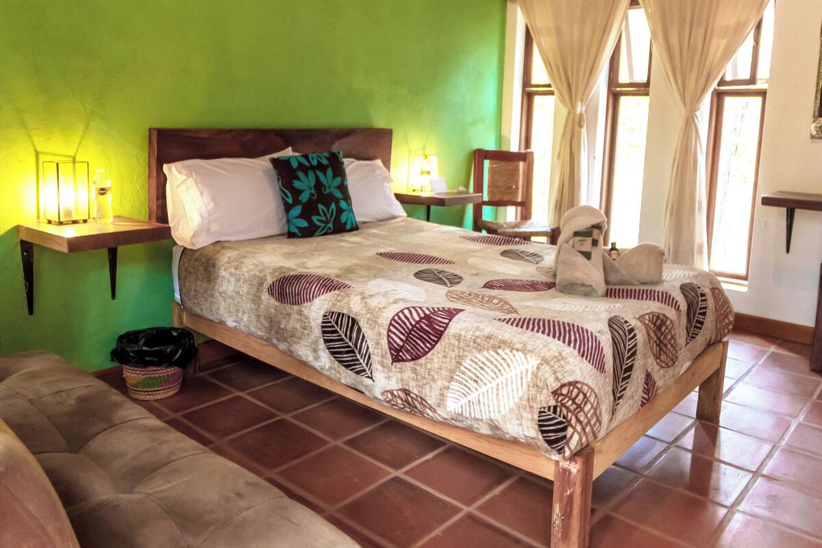 A bedroom with a large bed, green walls, wooden beams, a nightstand, a chair, and a window with curtains.