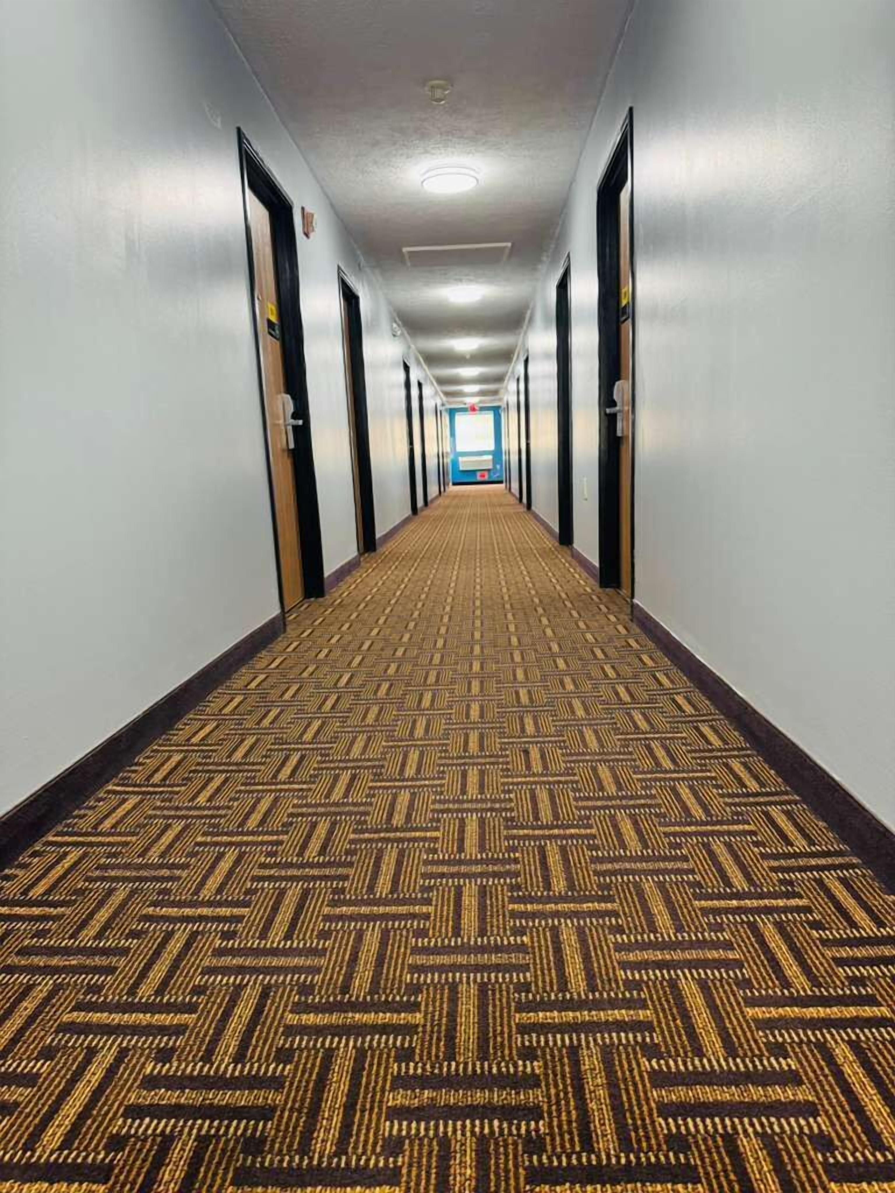 Un long couloir étroit, typique des hôtels, avec un revêtement de sol en moquette et des portes de chaque côté.