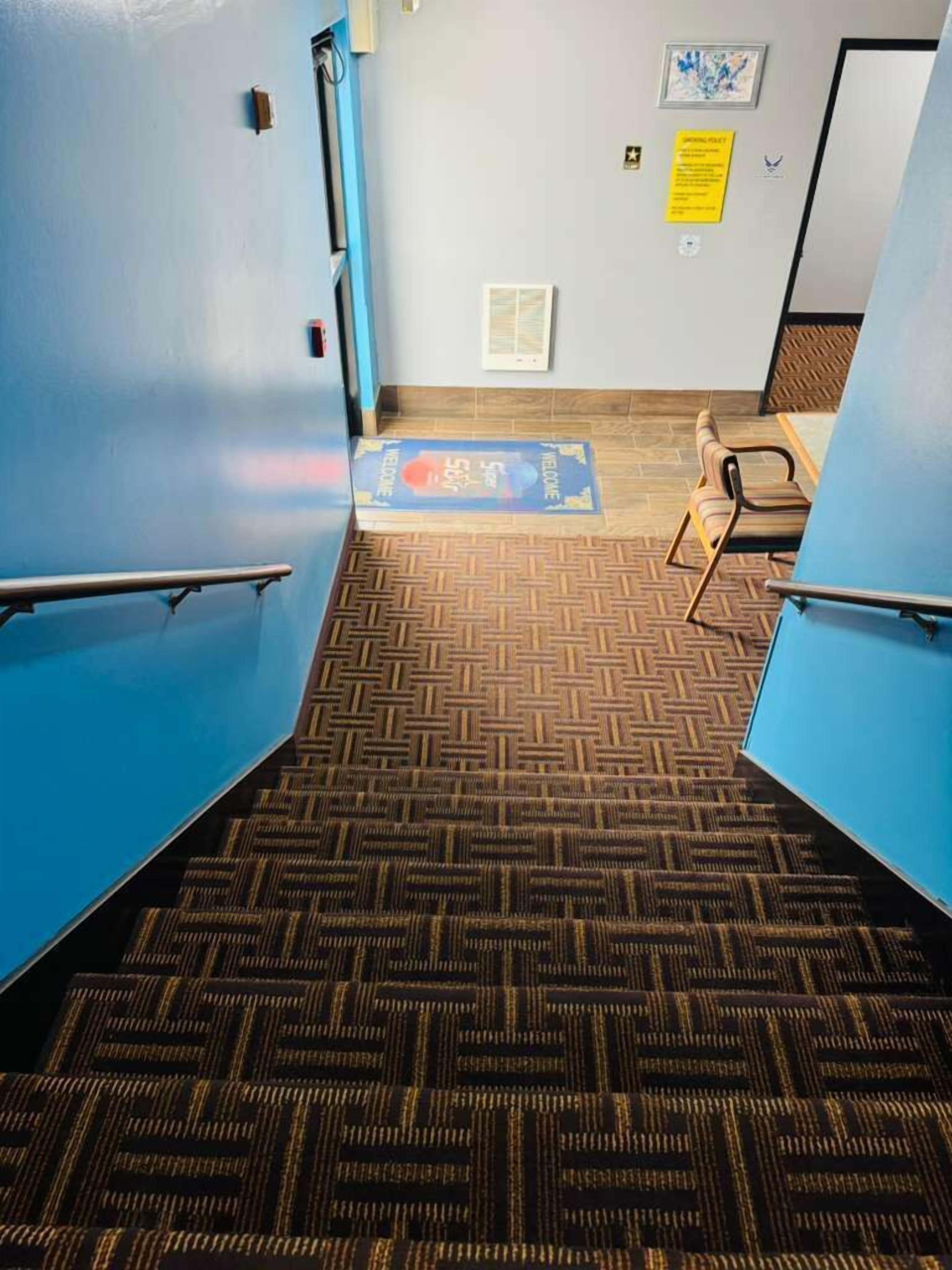 Una escalera alfombrada con un felpudo abajo, una puerta con un letrero y una pared con un cuadro enmarcado.