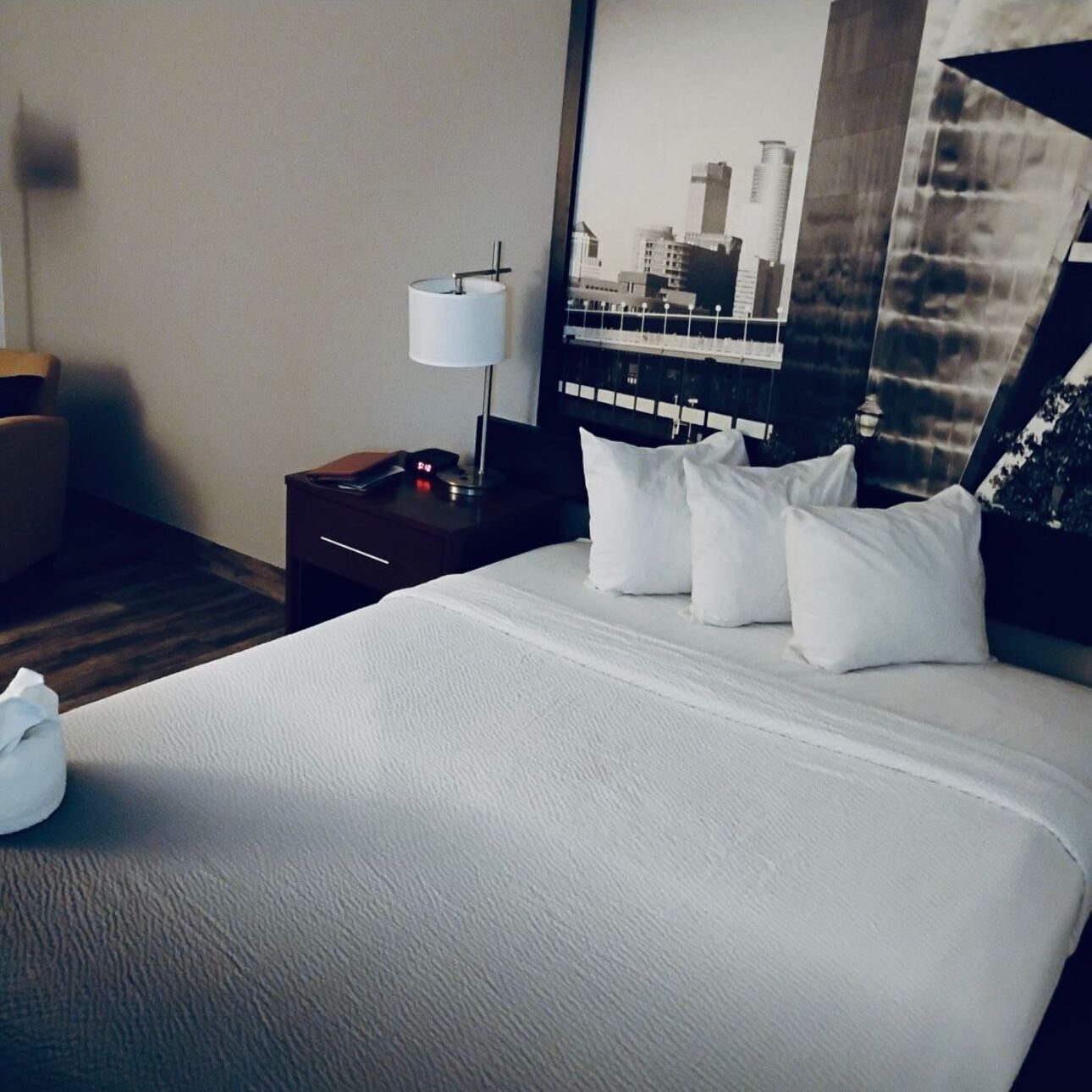 A hotel room with a large bed, a nightstand with a lamp, a clock, and a poster on the wall.