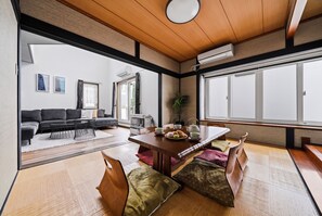 Ein traditioneller japanischer Raum mit Tatami-Boden, einem niedrigen Holztisch, der zum Essen gedeckt ist, einer Couch und Blick ins Freie durch große Fenster.