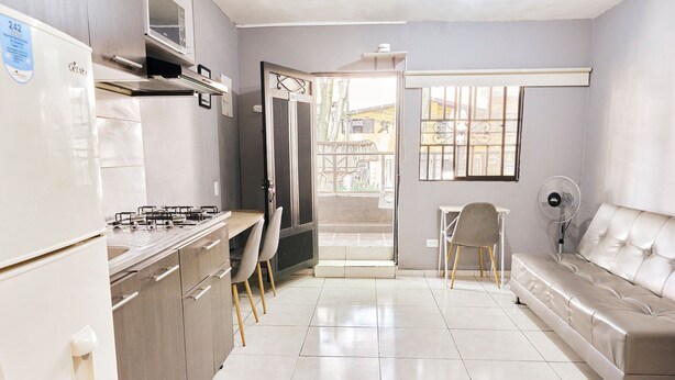 Une cuisine moderne équipée d’un réfrigérateur, d’une cuisinière et de chaises. Le salon comprend un canapé et un ventilateur.