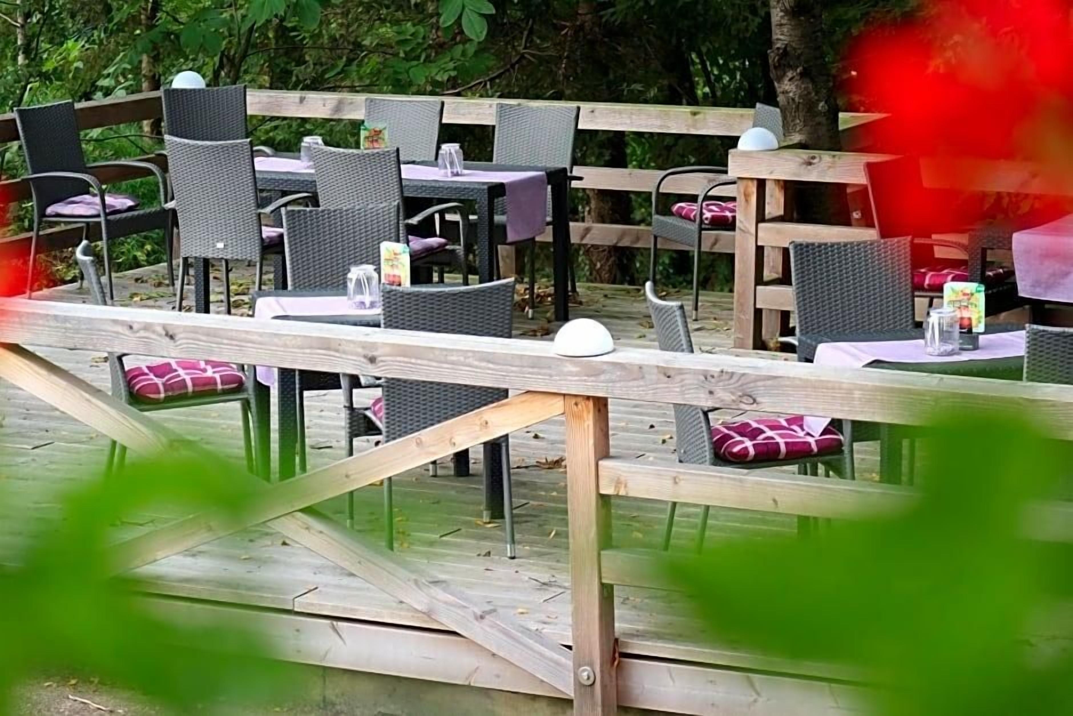 An outdoor dining area with wooden tables and chairs, surrounded by greenery.