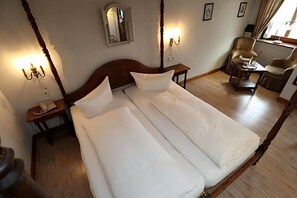 A double bed with white linens, a wooden headboard, bedside tables with lamps, and a chair with a table by a window. - Romantik Hotel Stollen (Gutach im Breisgau)