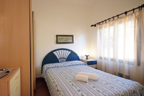 Une chambre à coucher avec un lit, une table de chevet, une lampe, un tableau encadré au mur et une fenêtre avec des rideaux.