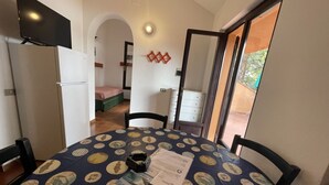 A dining area with a table set for two, a refrigerator, and a bed in the background. - Collina sul Golfo 13 (Porto Azzurro)