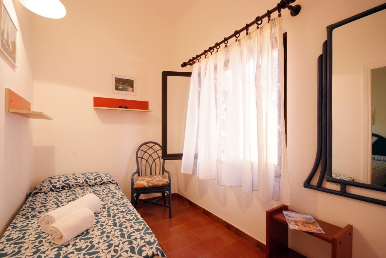 A bedroom with a bed, a chair, a shelf, a window with curtains, and a mirror.