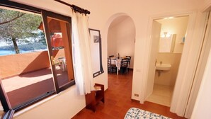 A room with a bed, a table with chairs, a bathroom, and a view of the outdoors. - Collina sul Golfo 13 (Porto Azzurro)