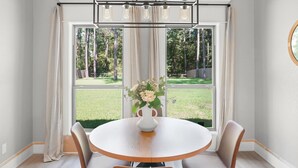 Uma área de refeições com uma mesa redonda, cadeiras, um vaso com flores e vista para um gramado através de grandes janelas.
