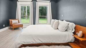 Quarto com cama grande, mesa de cabeceira de madeira, cadeira e duas janelas com cortinas.