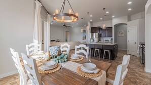 A dining area with a wooden table, white chairs, and a chandelier. There is a kitchen in the background with dark cabinets and a countertop. - 4BR Modern Farmhouse , Sleeps 10 , Backyard & Theater Room (Yuma)