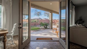 An open sliding glass door leading to a patio with a dining table and chairs, a small garden, and a view of neighboring houses. - 4BR Modern Farmhouse , Sleeps 10 , Backyard & Theater Room (Yuma)