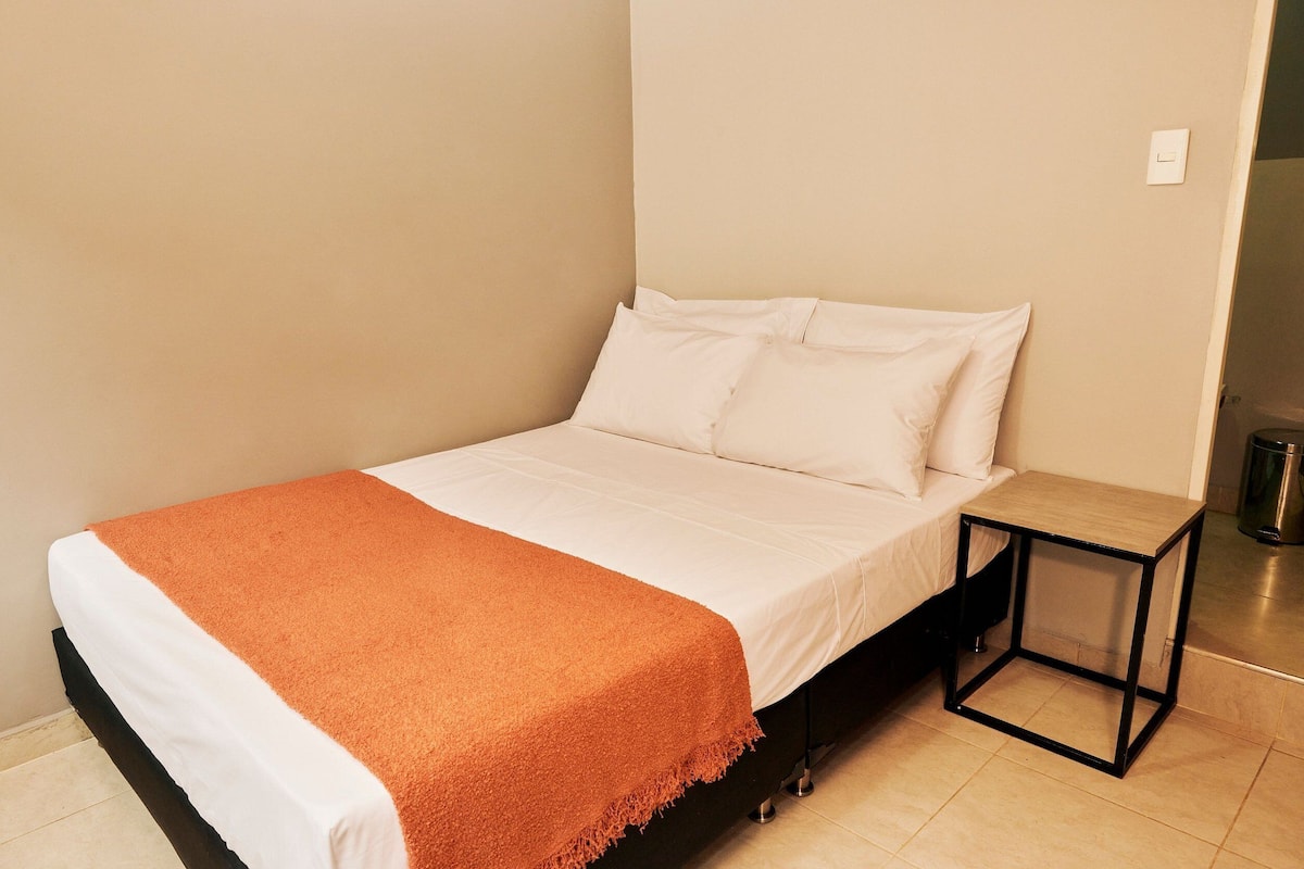 A neatly made bed with white linens and an orange throw blanket, a small bedside table, and a trash can in the corner.