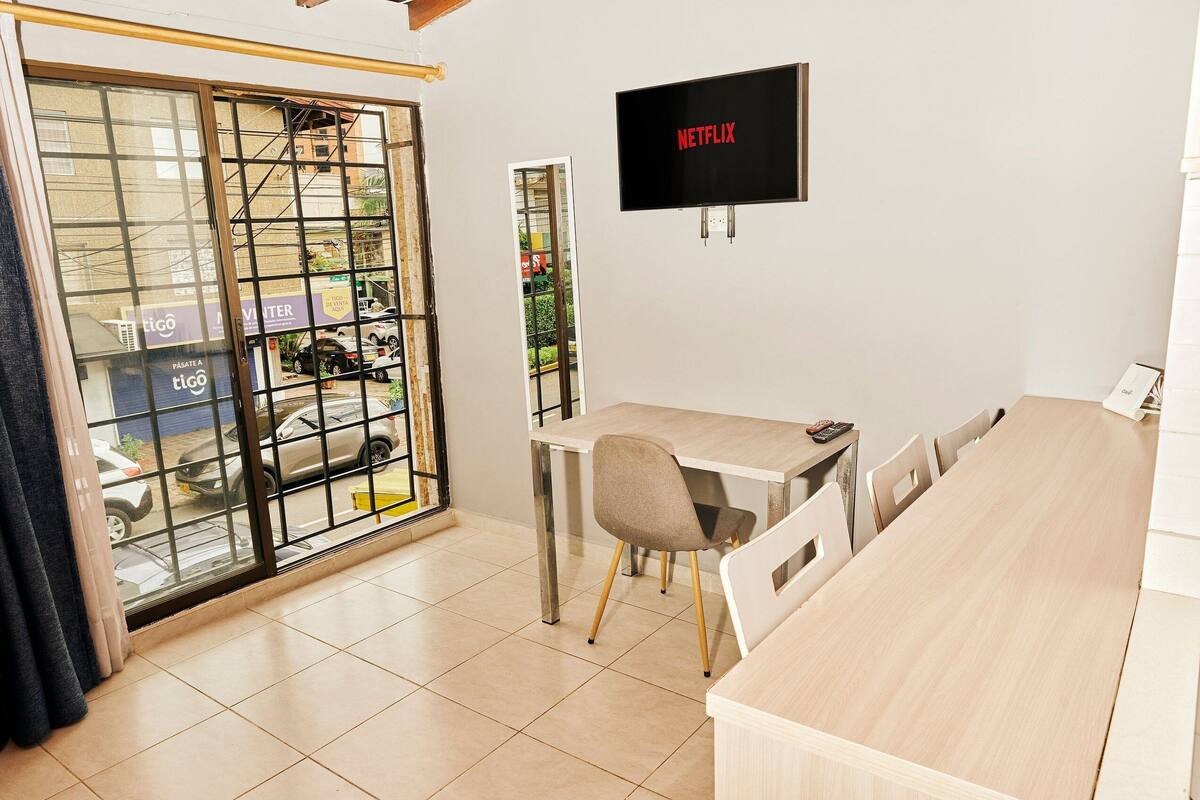 A modern room with a large window, a TV mounted on the wall, a wooden table, and a chair.