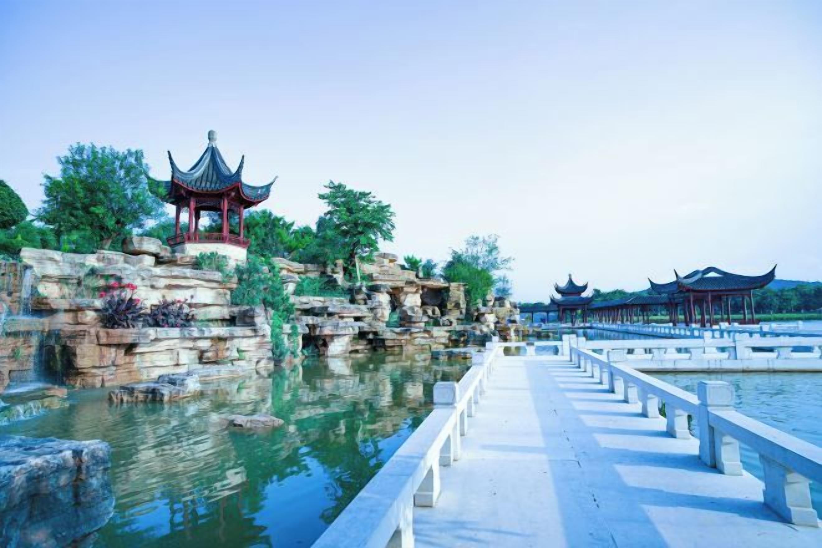 A traditional Chinese pavilion by a calm water body with a stone bridge leading to it.
