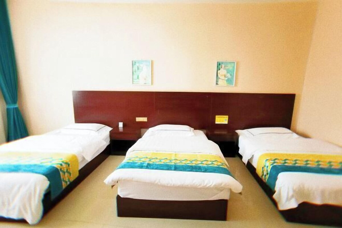 A hotel room with three single beds, each with a white mattress and a patterned bedspread. The room has a wooden headboard and two small wall art pieces above the beds.