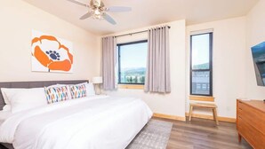 A bedroom with a large bed, a ceiling fan, a window with curtains, a wooden bench, and a TV on the wall.