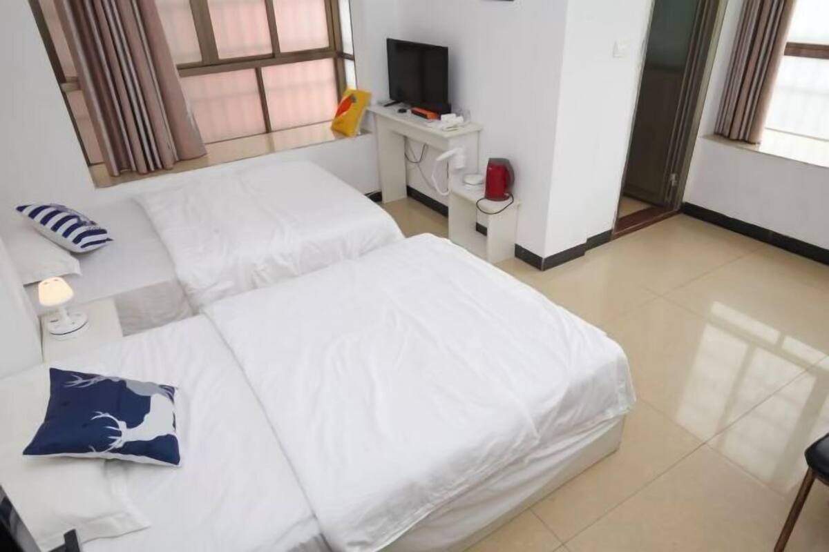 A hotel room with two beds, a desk with a TV, a red kettle, and a window with curtains.