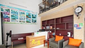A hotel reception area with a wooden reception desk, two grey sofas with orange cushions, a fan, and a clock on the wall. - Beihai Jiuyu Inn (Silver Beach) (Beihai)