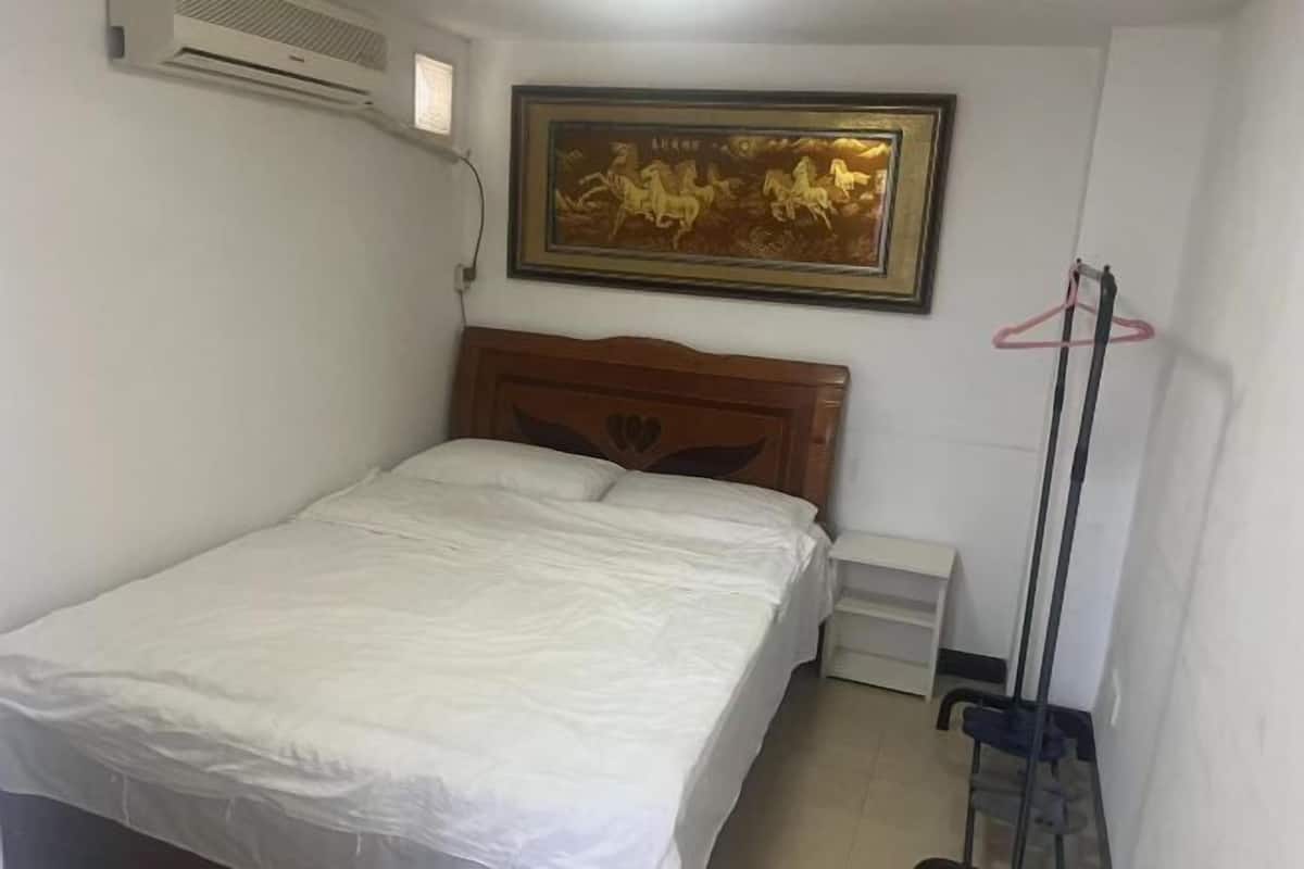 A bedroom with a bed, a framed picture, a white bedside table, and a wall-mounted air conditioner.