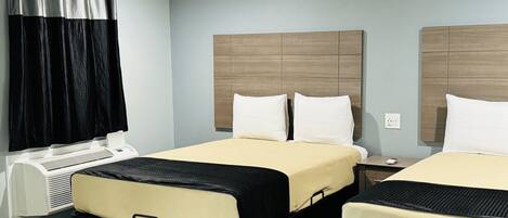 A hotel room with two beds, a large window with curtains, a bedside table with a lamp, and a wall-mounted air conditioning unit.