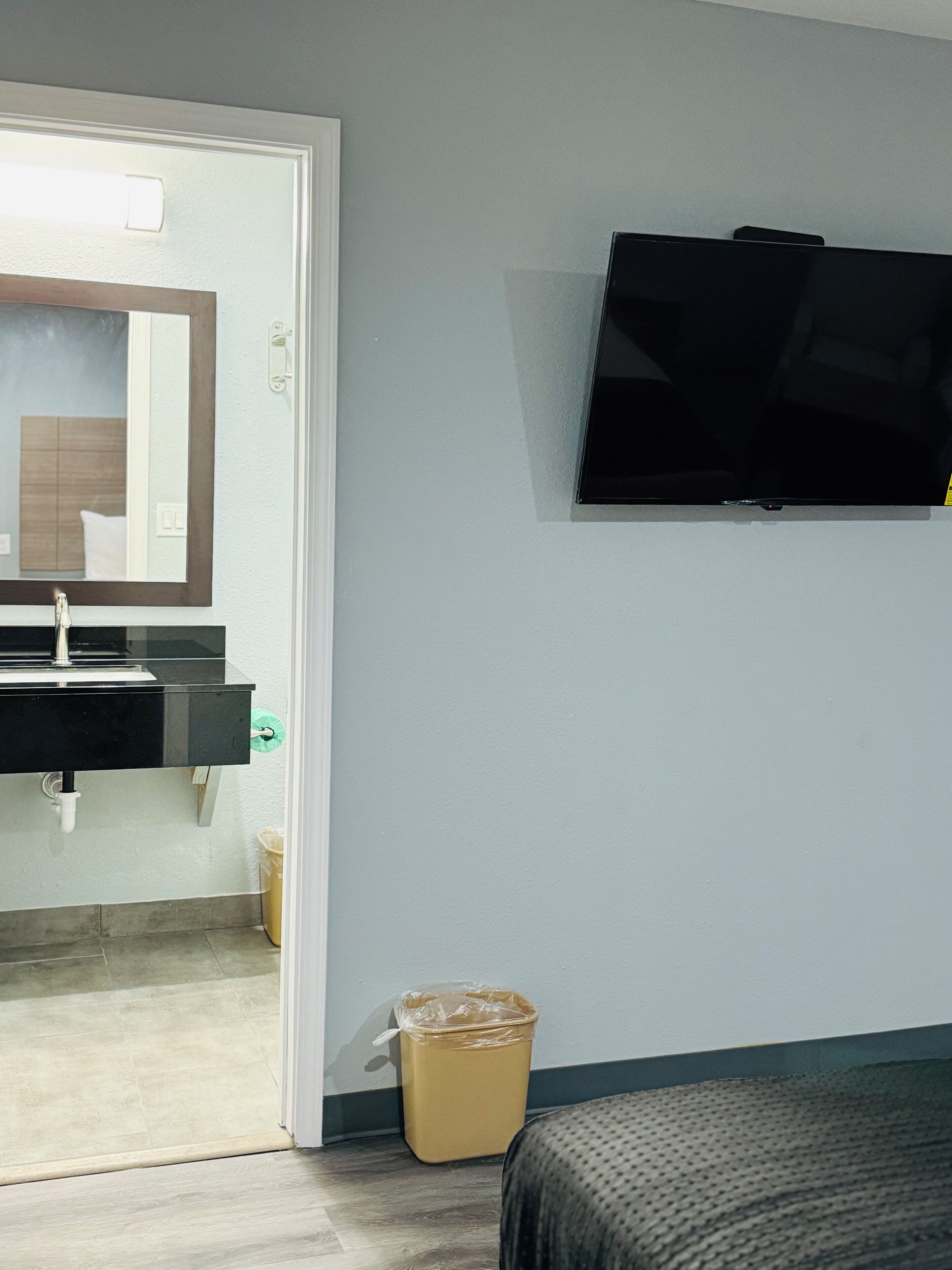 A hotel room with a bathroom, a TV mounted on the wall, and a trash bin.