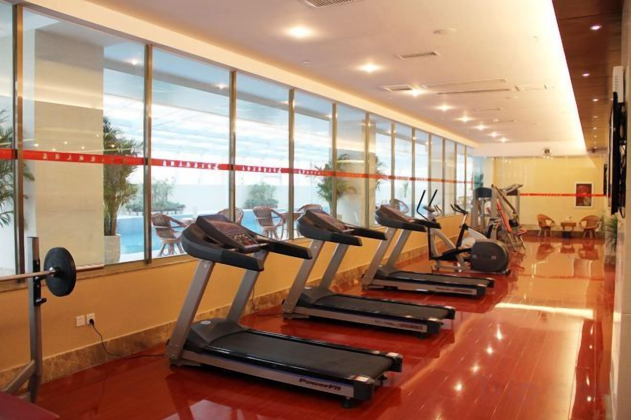 Un gimnasio con cintas de correr alineadas contra una pared con ventanas amplias, un banco para pesas y equipo de ejercicio.