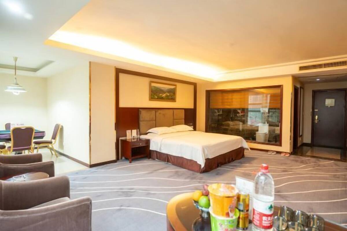 Habitación de hotel con cama, una mesa de comedor con sillas, una mesita con frutero y una botella de agua.