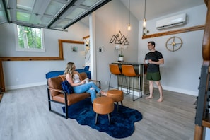 A modern interior with a sofa, ottomans, a dining table, and a person standing.