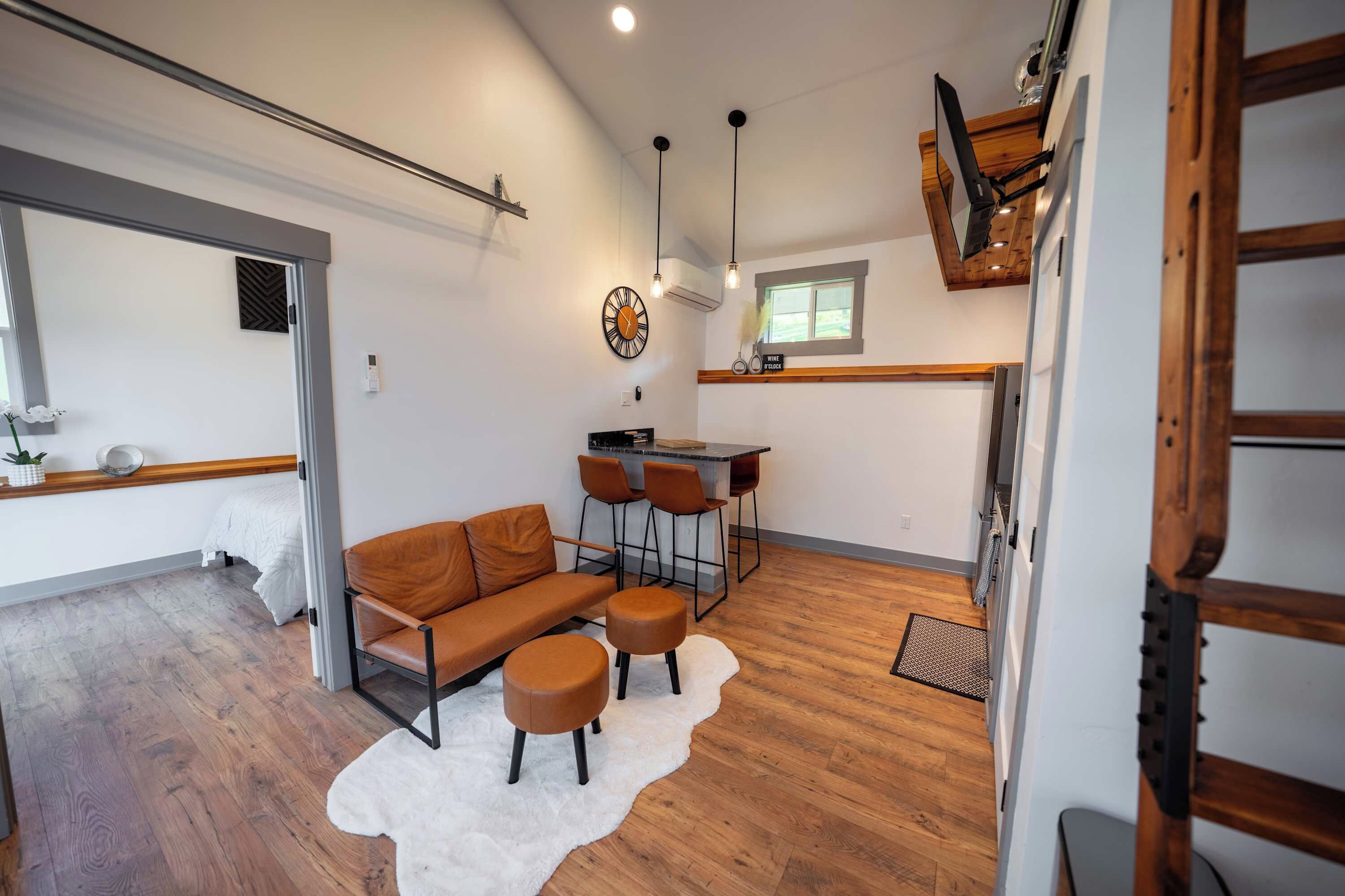 A modern living room with a sofa, ottomans, and a rug, a dining area with chairs, and a kitchen with a window.