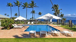 A pool area with lounge chairs and umbrellas, surrounded by palm trees and a view of the ocean.