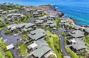 An aerial view of Kapalua Bay Villas 32B4, featuring a cluster of modern villas with sloped roofs, surrounded by lush greenery and a view of the ocean.