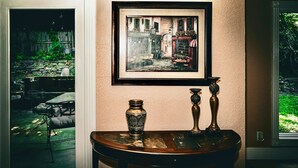 Ein gerahmtes Gemälde, das eine europäische Straßenszene zeigt, ein Holztisch mit einer Vase und Kerzenständern sowie der Blick auf einen Garten durch ein Fenster.