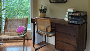 A room with a wooden desk, wicker chair, and a large window with a view of greenery.