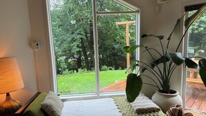 A room with a large window, a bed with a white bedspread and two pillows, a floor lamp, a potted plant, and a basket of towels.