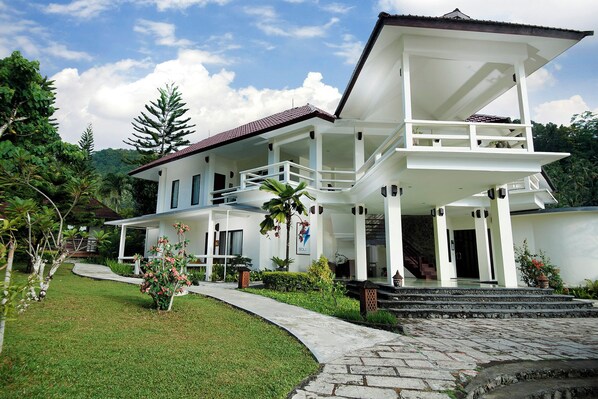 A two-story white building with a balcony, surrounded by greenery and a stone pathway.