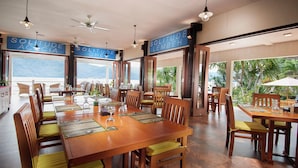 A dining area with wooden tables and chairs, large windows with ocean views, and ceiling fans. - Solitude Lembeh Resort (Bitung)